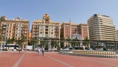 Mlaga, Andalusien Juli 26 2025: Mlaga 'da güneşli bir plaza, liman yakınlarındaki kentsel yaşamı gözler önüne seriyor. Yerel halk ve ziyaretçiler, ikonik AC Hotel Palacio da dahil olmak üzere modern otobüslerin ve tarihi binaların önünden geçtiler.. 