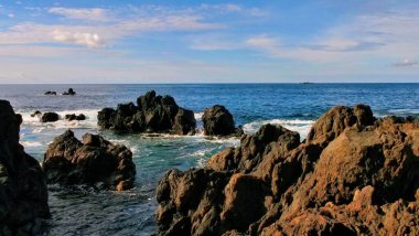 Çarpışan dalgalar, dağılmış bulutlarla dolu parlak bir gökyüzünün altındaki engebeli kıyı kayalarına çarptı. Dinamik sahne, evcilleştirilmemiş okyanus enerjisiyle dingin ufuk manzarasını harmanlıyor. Huşu ve doğal dramı çağrıştırıyor..