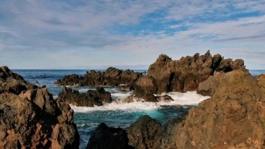 Çarpışan dalgalar, dağılmış bulutlarla dolu parlak bir gökyüzünün altındaki engebeli kıyı kayalarına çarptı. Dinamik sahne, evcilleştirilmemiş okyanus enerjisiyle dingin ufuk manzarasını harmanlıyor. Huşu ve doğal dramı çağrıştırıyor..