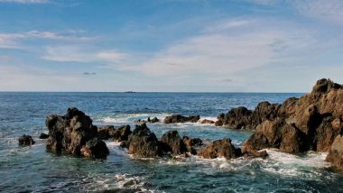 Karanlık kıyı kayaları ince bulutlu parlak bir gökyüzünün altında yavaşça dönen okyanus sularının ortasında duruyor. Tek bir kaya oluşumu ön planda hüküm sürüyor, güç ve zamansız yalnızlığı çağrıştırıyor..