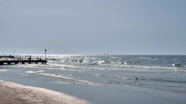 Yumuşak dalgalar sisli bir gökyüzünün altında sakin bir sahile yuvarlanıyor, güneş ışığı denizi parlaklaştırıyor ve uzaktaki bir iskele derinlik katıyor. Doğa, rahatlama ve kıyı seyahati temaları için ideal.