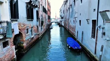 Eski binalar ve kırsal dokularla çevrili dar bir Venedik kanalında süzülen sürat teknesi. Uzak çan kulesi seyahat, şehir keşfi, denizcilik veya sinematik çekim için derinlik katar.