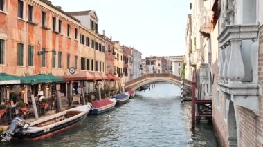 Klasik Venedik kanal sahnesi demirli tekneler ve yumuşak gün ışığında büyüleyici bir köprü. Seyahat reklamları, kültürel belgeseller veya Avrupa yaşam tarzı içeriği için mükemmel.