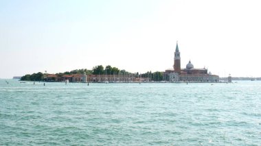 Sakin suları ve arkasında büyüleyici bir adası olan sakin bir sahil manzarası. Kırmızı kiremitli çatılar, küçük bir deniz feneri ve kenetlenmiş yelkenliler, açık mavi gökyüzünün altında Akdeniz dinginliğini uyandırıyor..