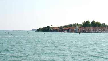 Sakin suları ve arkasında büyüleyici bir adası olan sakin bir sahil manzarası. Kırmızı kiremitli çatılar, küçük bir deniz feneri ve kenetlenmiş yelkenliler, açık mavi gökyüzünün altında Akdeniz dinginliğini uyandırıyor..