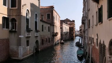Balkonları ve kemerli pencereleri olan binalarla çevrili Venedik kanalı. Tekneler durgun su boyunca yanaşıyor ve uzaktan küçük bir taş köprü geçiyor. Kanal kenarındaki hayata barışçıl bir bakış.