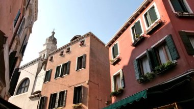 Venedik sokak sahnesi, sıva binalar, kemerli ve dikdörtgen pencereler, ve çiçek kutuları. Arka planda haç olan bir kilise kulesi, konut cazibesiyle kutsal mimariyi harmanlıyor.