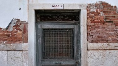 Yıpranmış yeşil kapı tuğla ve alçı duvarda, taş kemerle çerçevelenmiş. Alçı kabuğu altındaki kırmızı tuğlaları ortaya çıkarıyor. Metal bir ızgara pencereyi kaplıyor ve yukarıdaki tabelada Venedik yaşamına ait sessiz bir iz yazıyor..