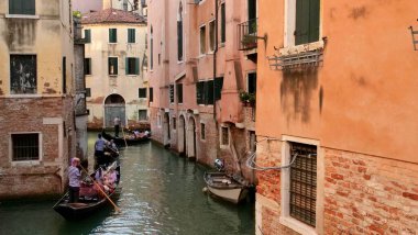 Çizgili tişörtlü gondolcuların kürek çektiği Venedik kanal sahnesi. Yolcular, yıpranmış balkonlu tuğlalı binalar ve zamansız cazibe ve hareket yakalayan yeşil fotoğraf makineleri arasındaki yolculuğun keyfini çıkarıyorlar..