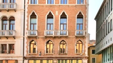 Pembe duvarları ve süslü beyaz taş takibi olan Venedik Gotik cephesi. Kemerli pencereler ve karmaşık detaylarla balkonlar simetrik, zarif bir kompozisyon oluşturarak zamansız mimari güzelliği yakalıyor..