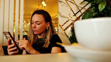 Kahvesini yudumlarken akıllı telefonuna odaklanmış rahat bir kafede oturan bir kadın. Ilık ışıklandırma, rahat bir ortam. Dijital yaşam tarzı, mobil kullanım, modern rutin veya kentsel kafe kültürü gibi temalar için ideal.