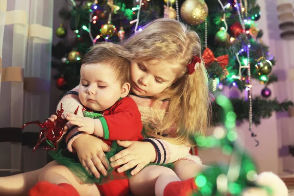 Two sisters are sitting by the Christmas tree, the older one hugs the younger one. The girls are playing together