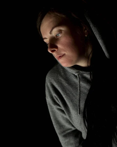 A girl in the dark, with her head covered. Black background behind the woman, a lamp shines on her from the side