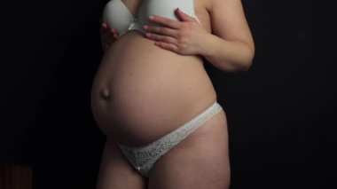 Pregnant woman in underwear. She is 30 weeks pregnant and has a big belly. Expecting a child. Third pregnancy. Pregnant girl. Black background and white underwear