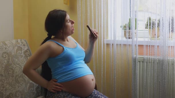 妊娠中の女性が窓のそばに座っている 子供を待ってる 女の子は妊娠中にチョコレートを食べる お菓子を欲しがる ダイエットに失敗します お腹が大きい 妊娠34週目 — ストック動画