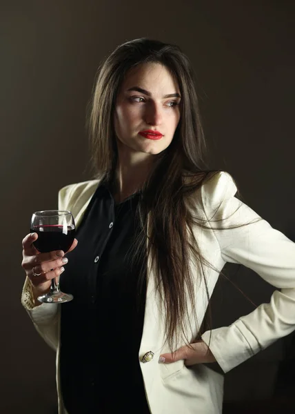 Şık giyinmiş güzel bir kız. Beyaz ceket ve siyah gömlek, stüdyo fotoğrafı. Model kameranın önünde poz veriyor. Elinde bir kadeh kırmızı şarap.