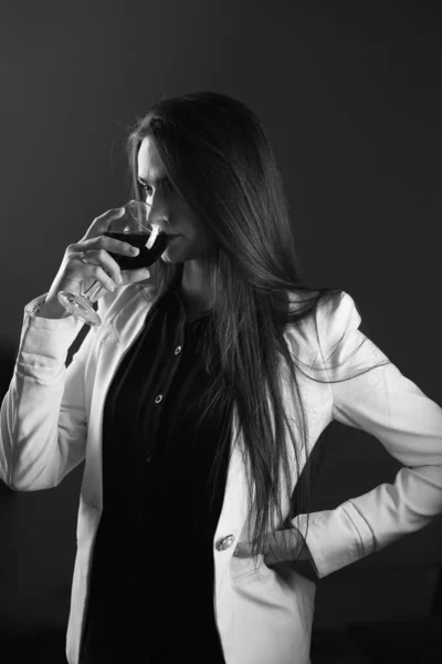 Beautiful girl in stylish clothes. White jacket and black shirt, studio shot. The model poses in front of the camera. Holding a glass of red wine