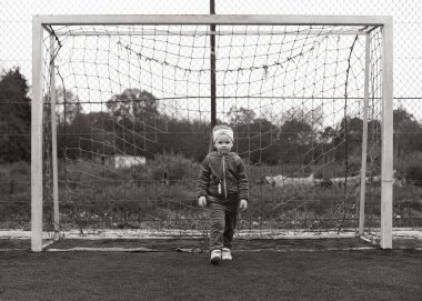 Küçük bir kız, 4 yaşında, kapının yanındaki bir tarlada duruyor. Bir çocuk futbol oynuyor. Kız yazın futbolu sever. Temiz havadaki çocukluk. Kız spora düşkün.