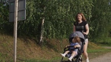 Bebek arabasında bir çocuk. Bir kız beşikte oturuyor, annesi el arabasını itiyor. Bebekle birlikte temiz havada bir yürüyüş. Gri bebek arabası, annelik, çocuk bakımı.