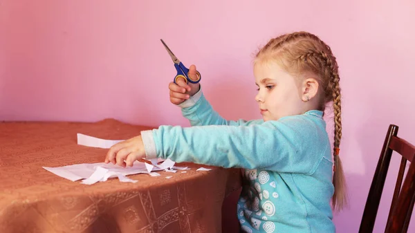 A child cuts paper with scissors. A little girl is sitting and cutting white paper. A preschooler learns to work with scissors. Program of preschool education at home. Child development, creativity