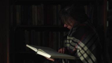 Bir kadın kitap okuyor, arka planda raflar ve birçok kitap var. Evde eğitim gören bir öğrenci evde edebiyat okuyor. Akıllı bir kız elinde bir kitap tutar. Gece atmosferi, geçmişe dönük yakın çekim.
