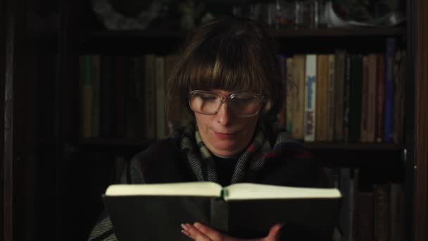 一个女人在看书 背后有很多书 他戴着滑稽眼镜 在家里学习文学 聪明的女孩手里拿着一本书 复古风格的特写 — 图库视频影像