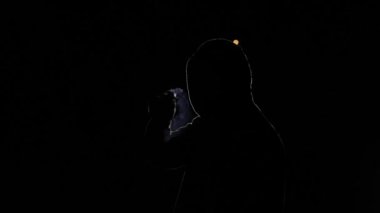 Siyah arka planda bir kişinin silueti. Kapüşonlu bir adam, ışıkla aydınlatılmış, patlar. Gecenin bir hackerı, yüzü görünmeyen gizemli bir insan. Bir sigara içiyor.