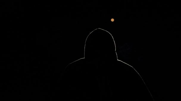 黒い背景の人のシルエット 光に照らされたフードの中の男 夜のハッカー その顔が見えない神秘的な人物 タバコを吸っている — ストック動画