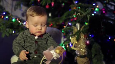 Çocuk Noel ağacının yanında oturuyor. Şenlikli atmosfer, yeni yıl ve Noel ağacı. Küçük çocuk güzel giyimli ve sarı saçlı. Çocuklar için Noel, mutlu yıllar.