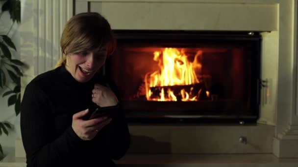 靠近壁炉的那个女孩 手里拿着电话 一个女人坐在网上 一个火在后面的壁炉里燃烧 舒适的气氛 圣诞前一晚 — 图库视频影像