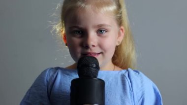 Küçük bir kız mikrofon tutarken şarkı söylüyor. Sarışın, mavi takım elbiseli güzel bir aktris. Çocuklar için karaoke, genç şarkıcı
