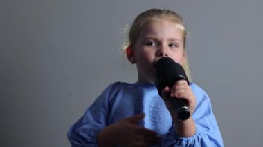 Küçük bir kız mikrofon tutarken şarkı söylüyor. Sarışın, mavi takım elbiseli güzel bir aktris. Çocuklar için karaoke, genç şarkıcı