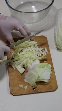 Eldiven giyerken tahta kesme tahtasının üzerinde taze lahana doğrayan biri. Bir kase su yakınlarda oturur, temizliği ve yemek pişirme hazırlıklarını vurgular..
