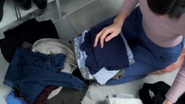 Kadın elbiselerini düzgünce katlayarak dolabını düzenliyor. O, yerde oturuyor ve elbiselerini düzenliyor. Ev hanımı soyunma odasındaki dolabı topluyor..