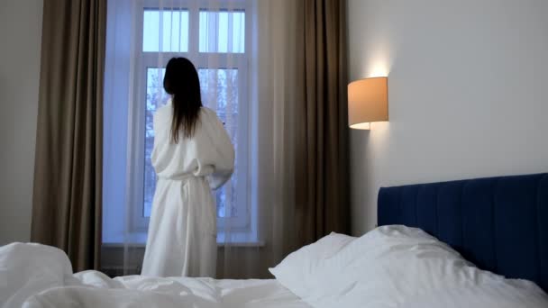 穿着白色浴衣的布鲁内特女人们正在观望着酒店房间的窗户上的寒假 回头看 带着舒适的旅行概念 — 图库视频影像