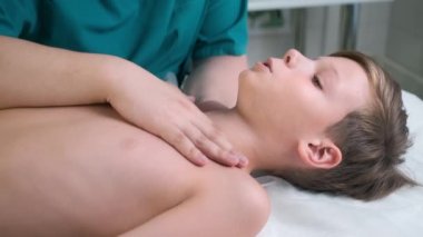 Kraniosakral terapi seansı, ergen erkek omurgasının bir doktor tarafından tedavisi. Çocuk kanepede yatıyor. Kraniosakral terapist, omurga duruşunu düzeltmek ve iyileştirmek için köprücük kemiğine ve omuzlarına dokunuyor..