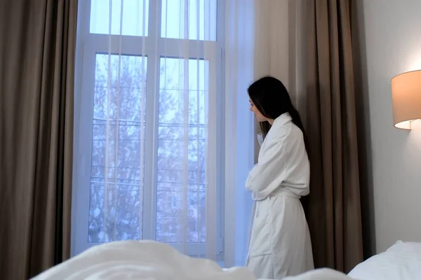 白いバスローブの若いスリムな女性がホテルの部屋の冬の木の窓を見ています 彼女は笑顔でベッドの近くに立っている 冬のコンセプトで快適さ リラックスして休息 — ストック写真