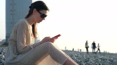 Batumi şehri Georgia 'da çakıl taşlı deniz kıyısında oturan akıllı telefonlara bakan bir kadın. Sosyal medyada telefonda kadın sohbeti. Yaz tatili, plaj gezisi deniz konseptine seyahat.