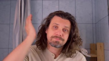 Banyoda yüzüne dokunan yorgun sakallı adam aynada görünüşünü şok etti. Kıvırcık uzun saçlı adam stres, uykusuzluk, depresyon ve anksiyete yüzünden acı çekiyor. Uykulu üzgün erkek.