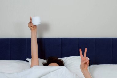Battaniyenin altında saklanan esmer kadın zafer işareti gösteriyor ve elinde bir fincan kahveyle yatakta yatıyor. Tatil, tembel sabah, dinlenme ve rahatlama konsepti. Uyanıp çayını içmeye çalışıyor..