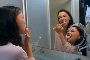 Esmer, orta yaşlı bir kadın banyoda aynaya bakarken plastik diş fırçası kullanıyor. Kadın sabah yeni bir güne hazırlanıyor. Evde diş temizliğine önem veriyordu. Diş bakımı.