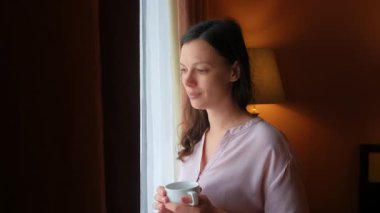 Esmer kadın çay içiyor pencereye bakıyor gülümsüyor ve eğleniyor. Kadın kahvaltı yapıyor ve işe hazırlanıyor. İyimser orta yaşlı insan tek başına taze içecek içer..