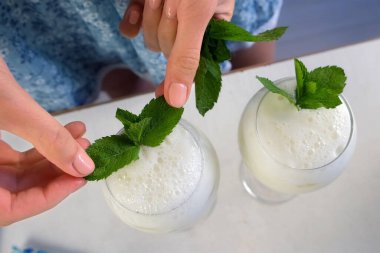 Beyaz Brezilya limonatasını şarap bardaklarıyla süsleyen bir kadın. Mutfakta nane yaprağı, üst manzara. Yemek tarifi, profesyonel aşçılık. Turunçgil meyveli ev yapımı içecek hazırla..
