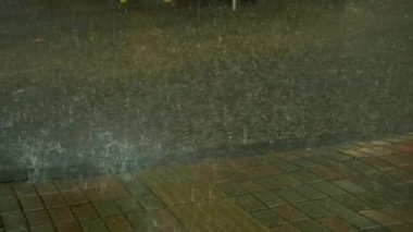 Heavy rain falling on city street asphalt and pavement creating huge puddles. Rainwater cascading over tiled sidewalk and asphalt street during heavy nighttime urban downpour.