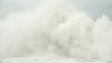 Powerful sea foamy waves violently breaking against rocky shoreline, revealing dramatic natural forces during cloudy overcast weather. Giant ocean waves crashing on pebble shore in stormy weather.