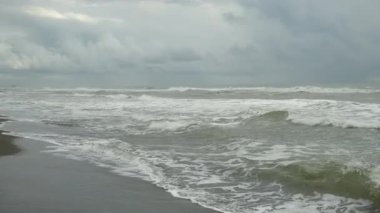 Stormy sea waves rolling crashing on black volcanic sandy beach under cloudy sky. Creating dramatic powerful scene. Beauty of nature in overcast weather. Wonderful seascape from coastline.