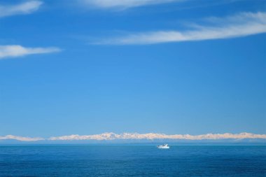 Deniz kıyısında karlı dağları ve limandan yüzen beyaz yatı olan doğal mavi bir arka plan. Turistik deniz teknede yürüyor, gemi güneşli kış havasında. Turizm, seyahat, tatil, seyahat kavramı.