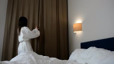 Neşeli esmer kadın sabah otel odasında perdeleri açıyor. O, tatilinde bir kış gününe başlıyor. TRavel, yolculuk, turizm, karlı orman manzaralı rahat bir otelde dinlenme..