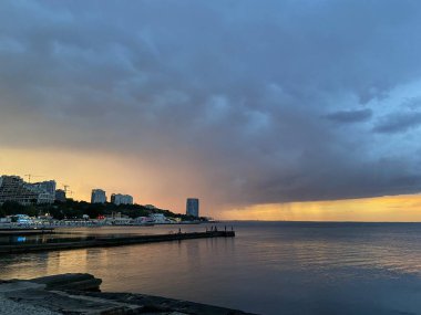 Büyük şehrin kıyı bölgelerinin akşam ışıkları ve batan güneşin son ışınlarıyla aydınlanan gökdelenlerin üzerinde asılı ağır gri bulutların inanılmaz bir kombinasyonu..