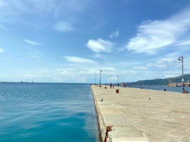 Adriyatik kıyılarının sakin su yüzeyinin panoraması, Trieste şehrinin gezinti güvertesine yakın, ancak bulutlu mavi bir gökyüzünün arka planına karşı..
